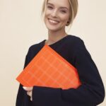 A cheerful woman smiles while holding an orange folder against a neutral background.